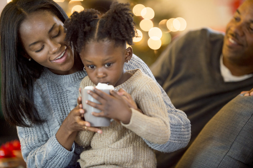 A little girl is drinking hot cocoa with her parents on Christmas morning in their home.