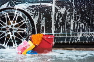 Bucket with sponges and brush with lots of foam next to car tire.